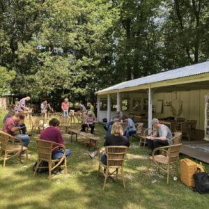 Een groep mensen die deelnemen aan de UITVERKOCHT Magische Mantra Basis Workshop, zittend rond een tafel in De Hoorneboeg 't Gooi, een bosrijke omgeving.